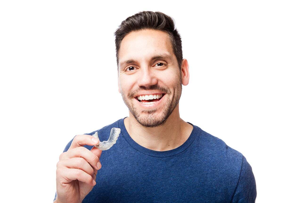 Man holding clear Invisalign aligner, smiling at camera in dental office setting.