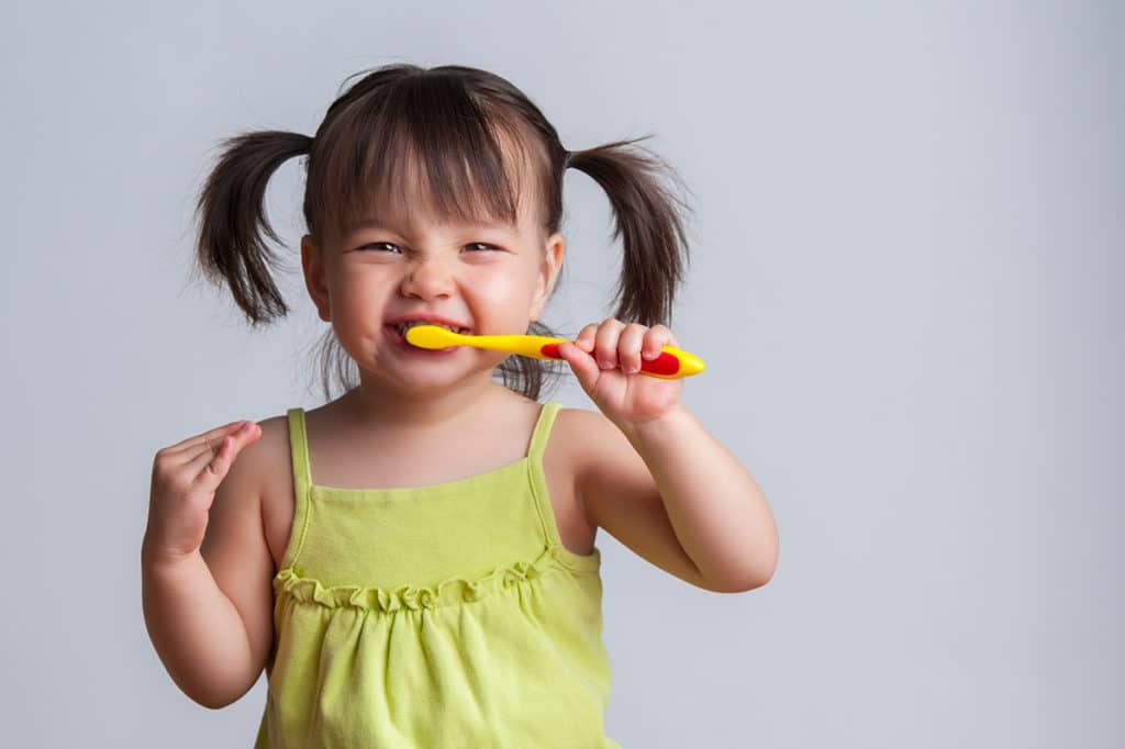 Smiling toddler with pigtails brushing teeth with a yellow and red toothbrush.
