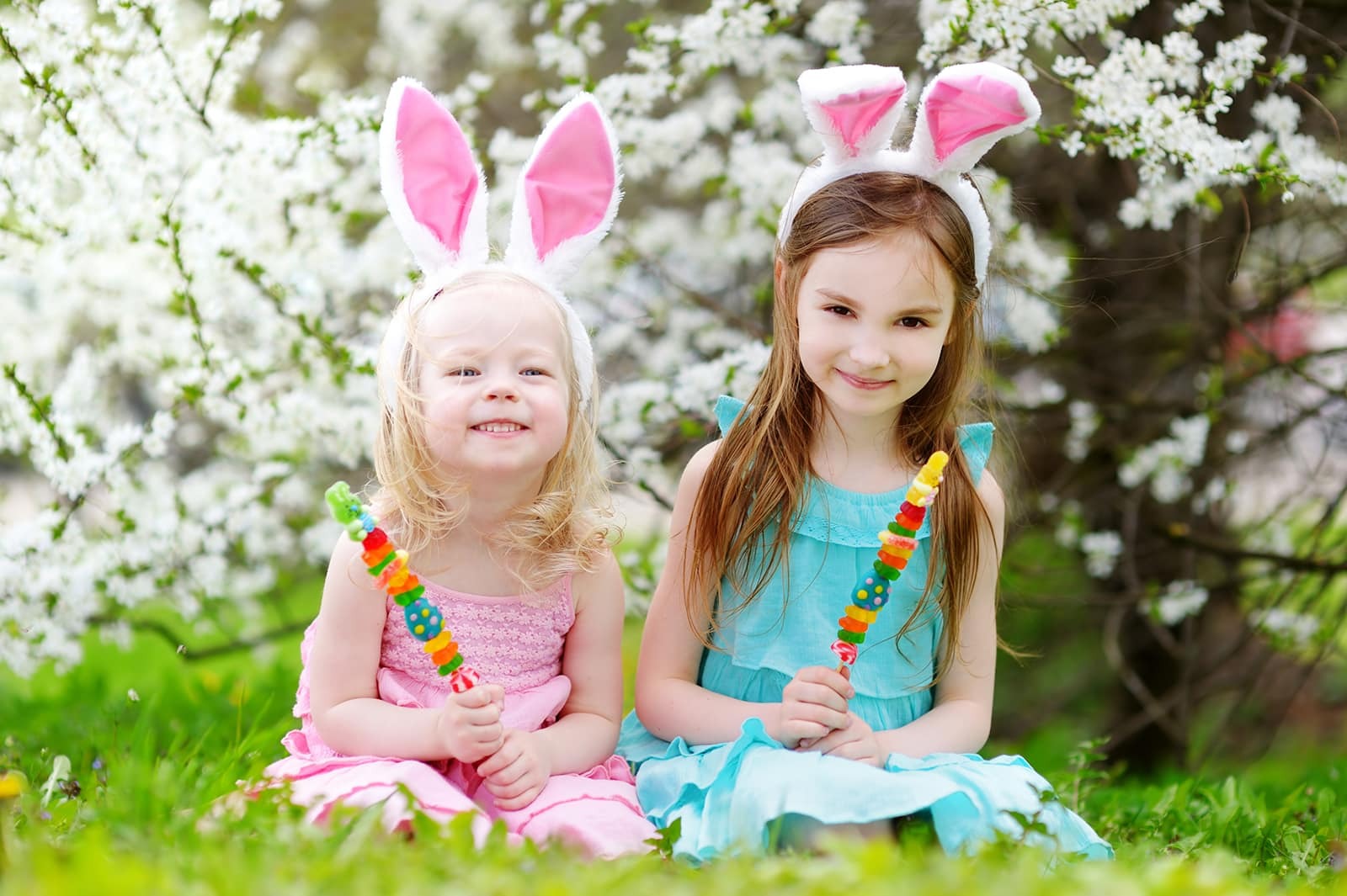 Two young girls wearing bunny ears smile while holding colorful candy near blooming white flowers.
