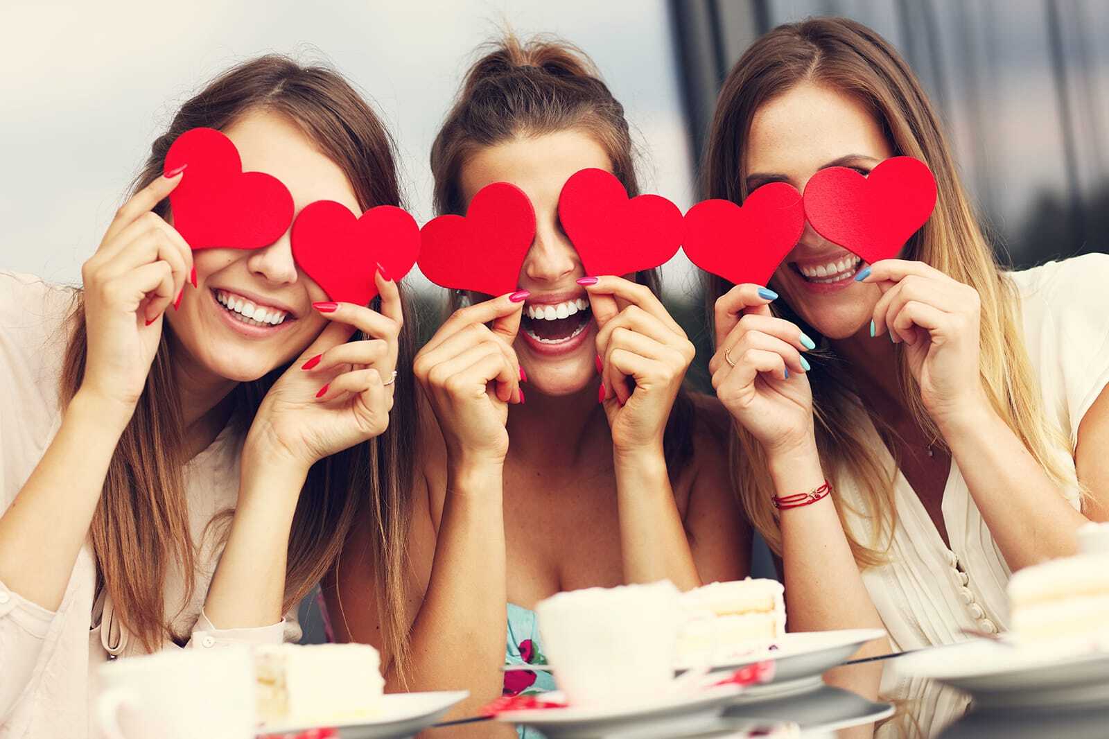 Three smiling women holding red hearts together at a cafe table.