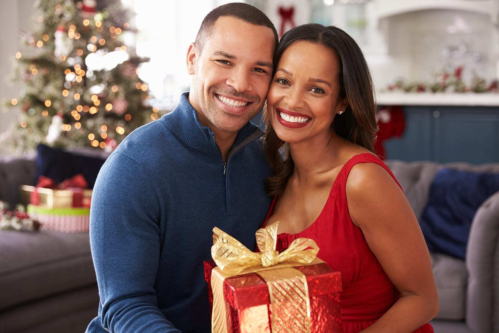 Smiling couple holding a wrapped gift in a festive holiday living room setting.