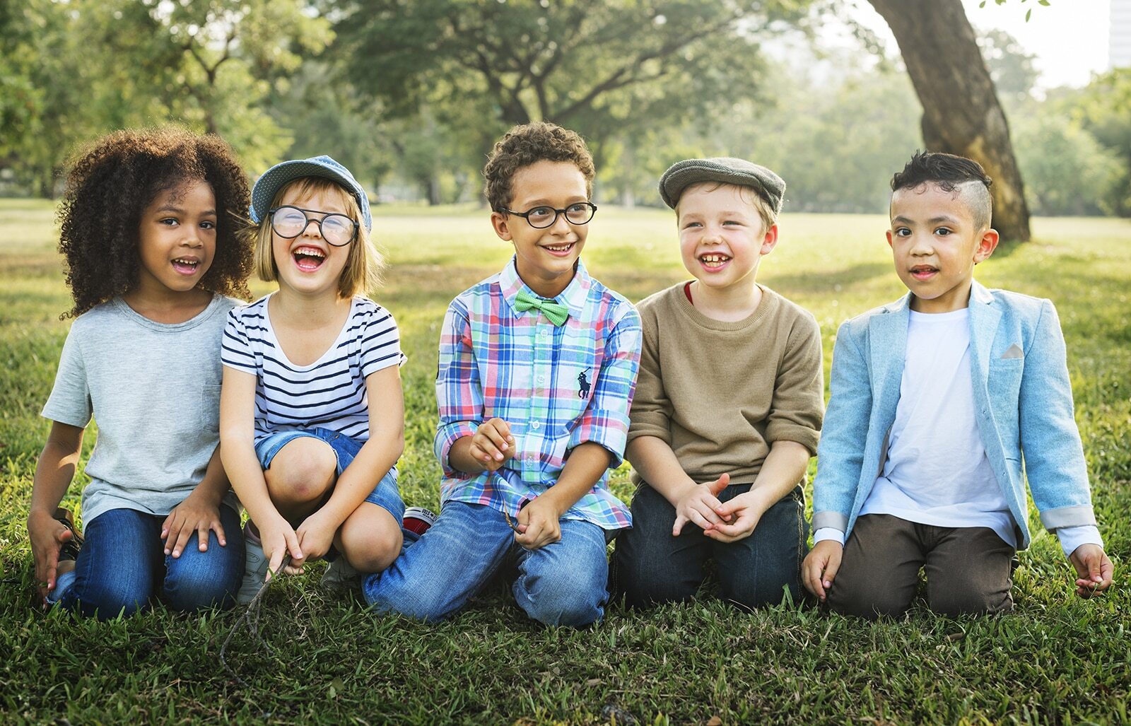 Five smiling children sitting together on grass in a park setting.