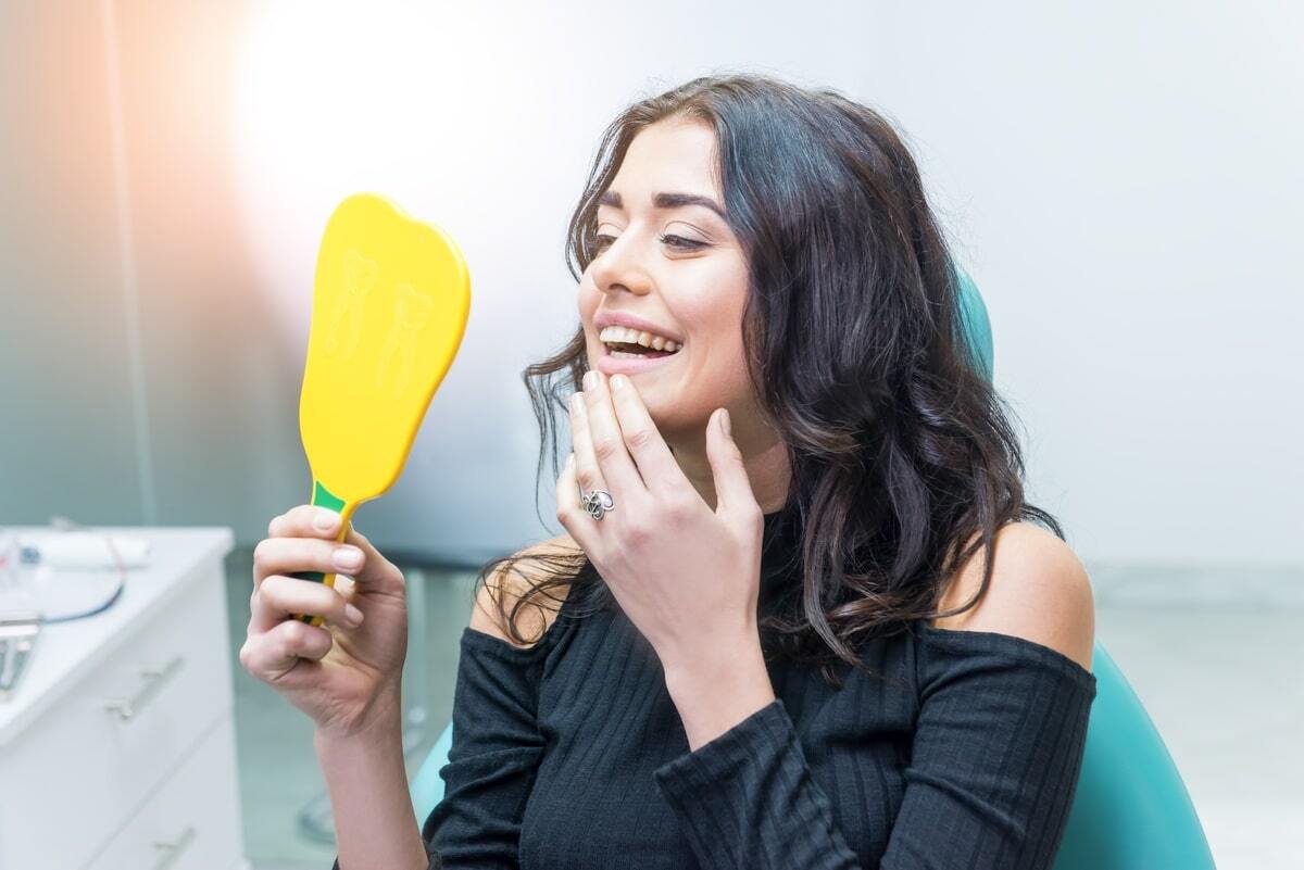 Woman examining her smile in a dental mirror during a consultation about tooth replacement options.