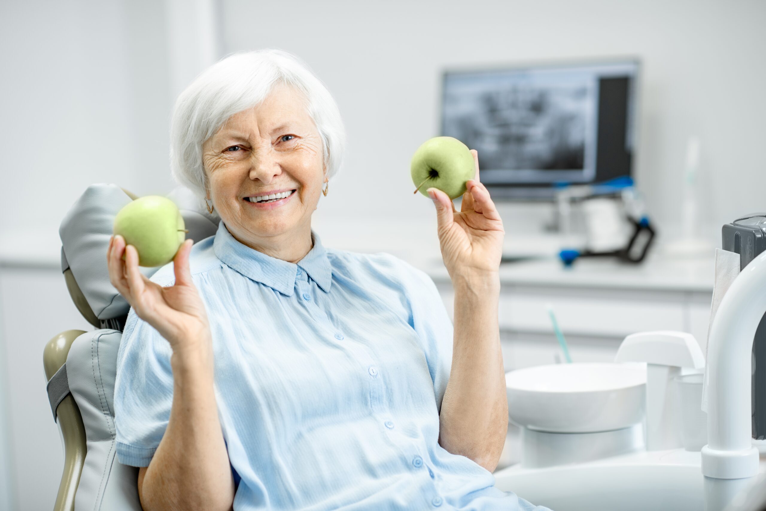 Senior woman holding green apples in a dental office, demonstrating healthy food choices for dental implant care.