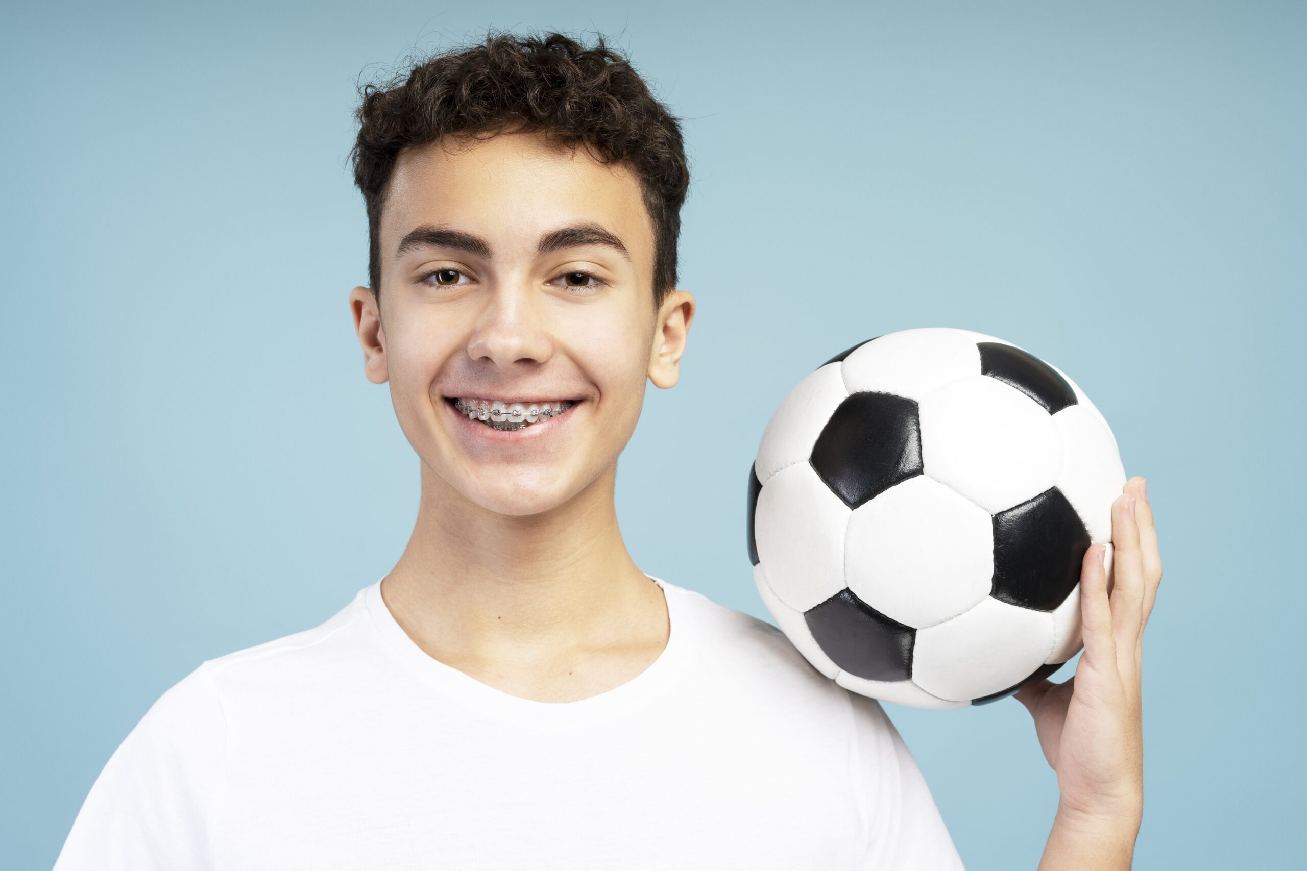 Teenage boy with braces smiling while holding a soccer ball against blue background.