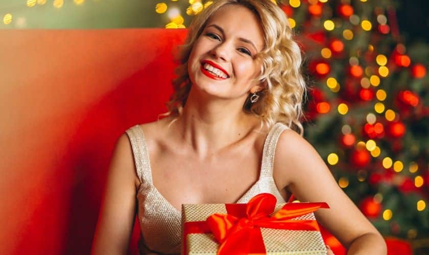 Woman smiling confidently while holding a wrapped gift, surrounded by festive holiday lights.