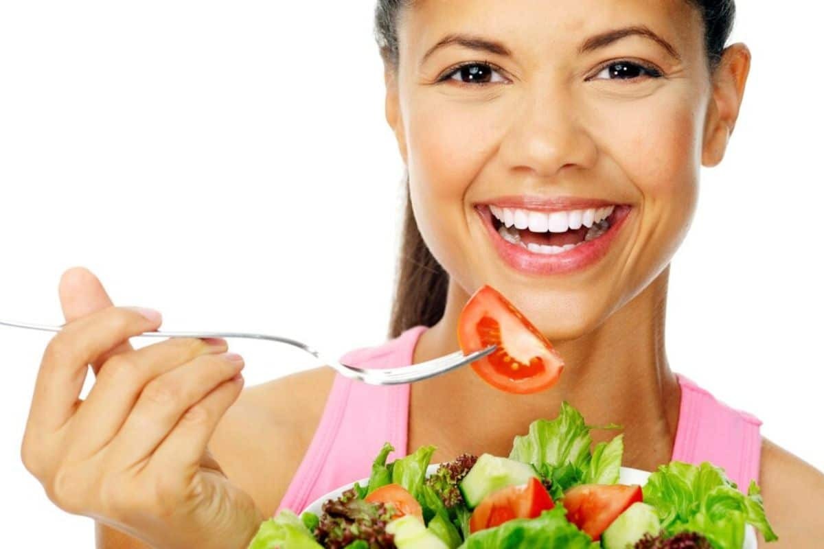 Woman in pink top eating healthy salad with tomato and lettuce, smiling with white teeth.