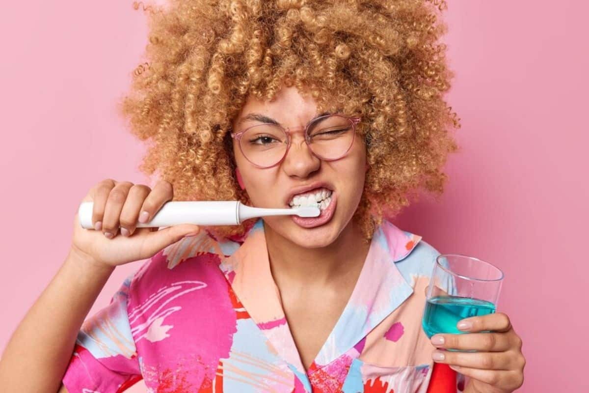Woman brushing teeth with electric toothbrush and mouthwash, demonstrating proper oral hygiene practices.