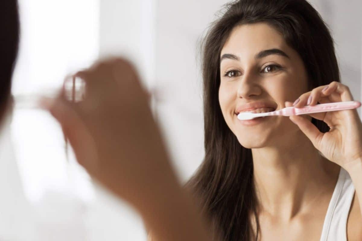 Woman brushing her teeth with a pink toothbrush and smiling at the mirror.