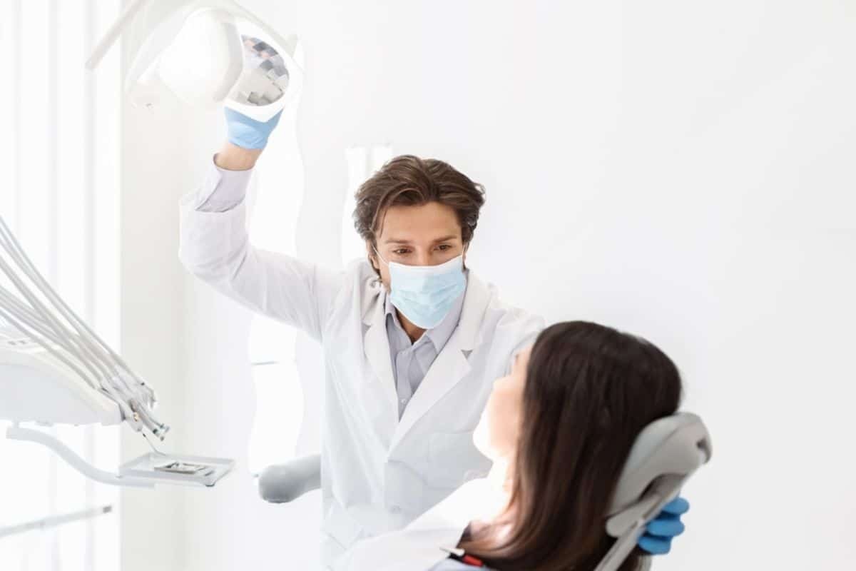 Dentist in white coat and mask adjusting overhead light during patient examination in bright dental office.