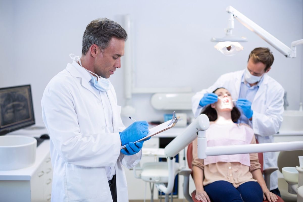 Dentist reviewing patient records while colleague examines patient in Corpus Christi dental office.