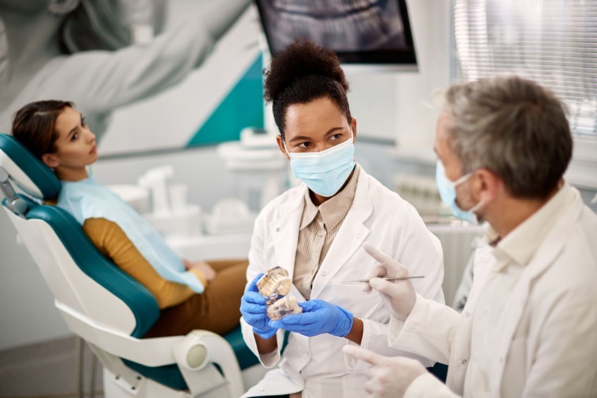 Female dentist discussing dental model with male colleague while patient sits in dental chair.