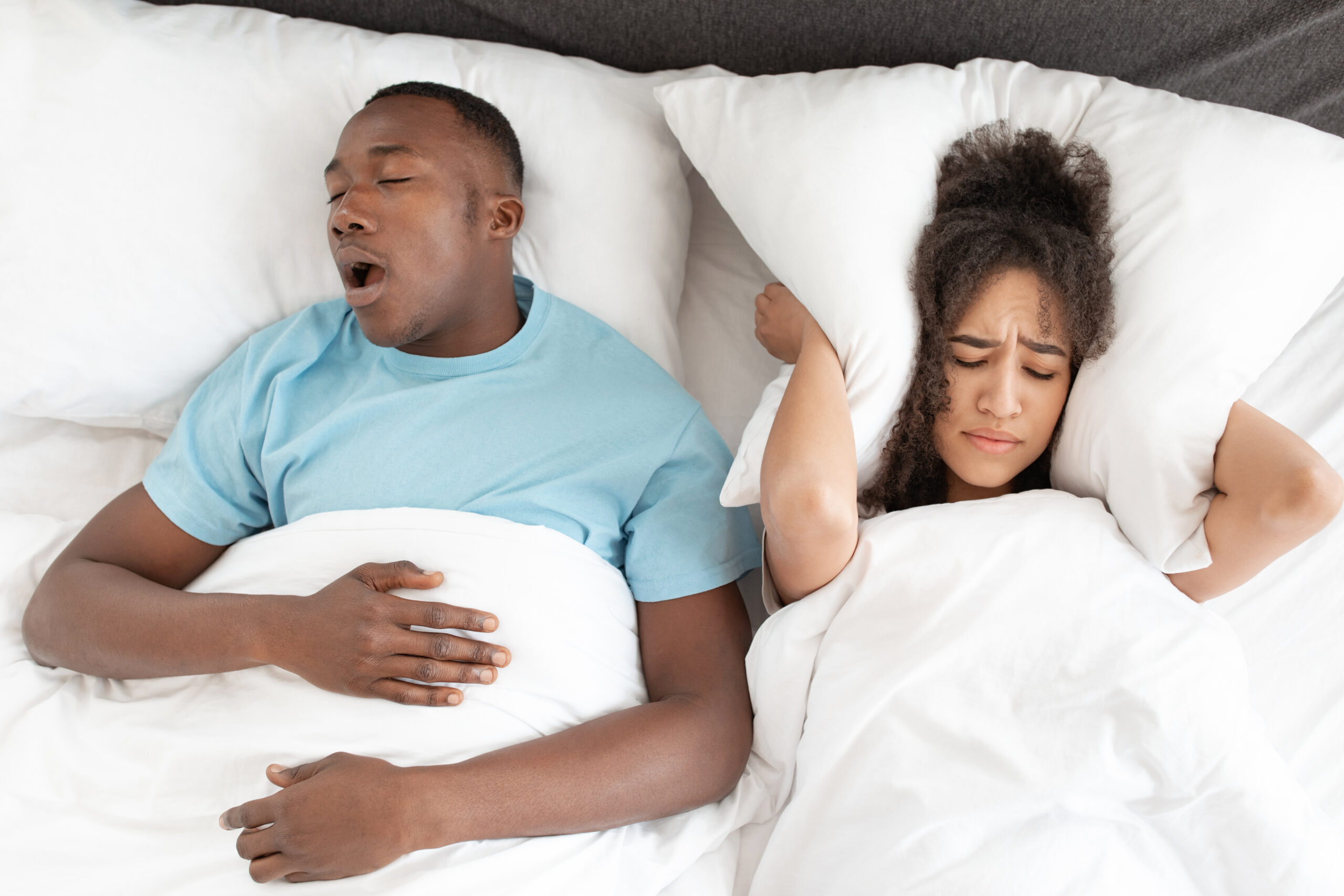 Man snoring in bed while woman covers ears, illustrating sleep disruption related to snoring issues.