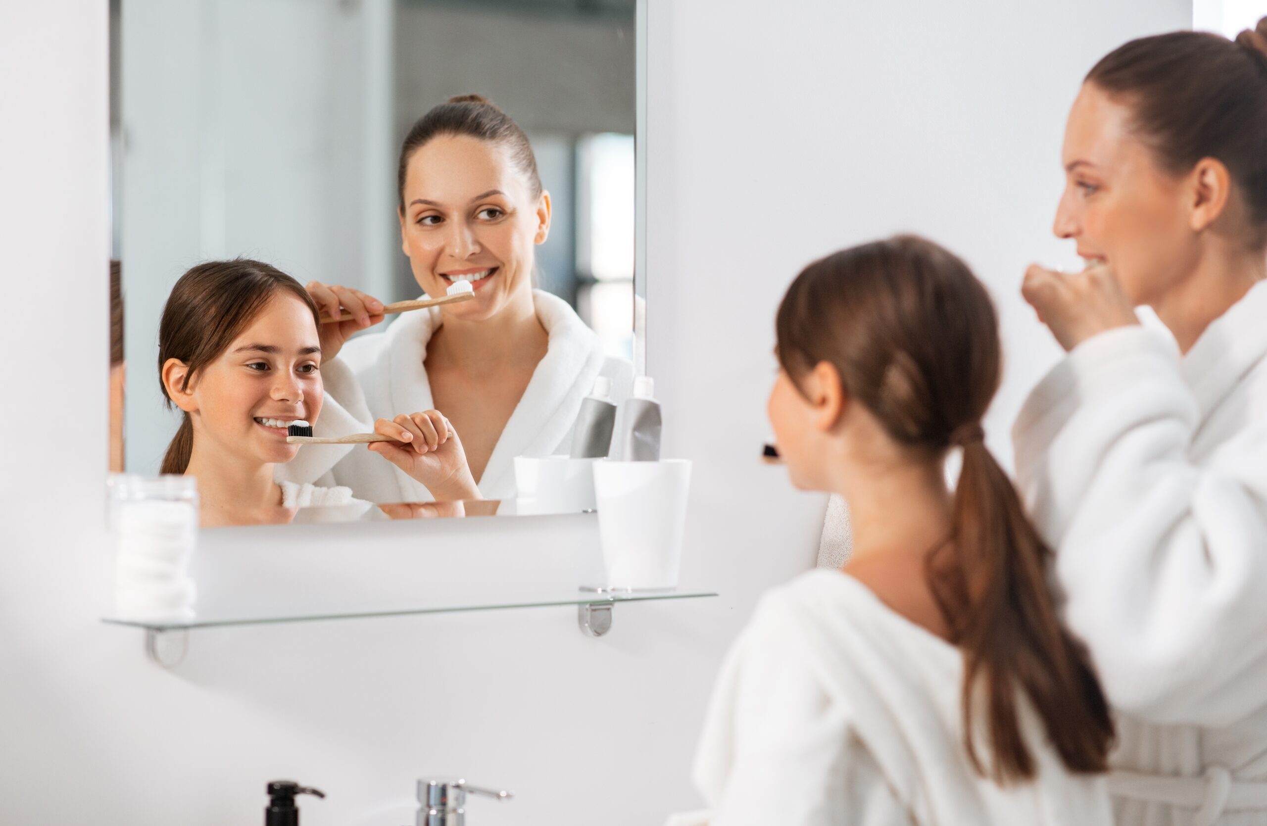 Family brushing teeth together in bathroom mirror, demonstrating daily oral hygiene habits.