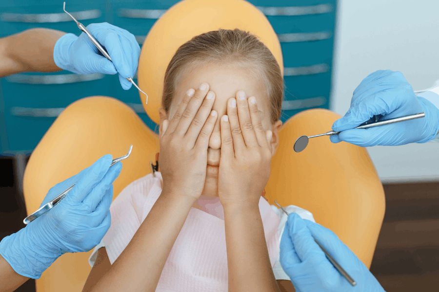 Child covering face during dental visit, surrounded by dentist tools and yellow dental chair.