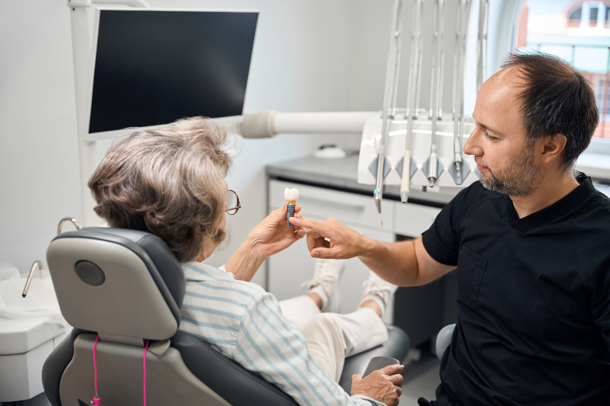 Dentist showing dental implant model to senior patient in treatment room at special needs dental practice.